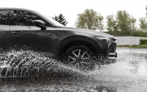 Ciemny samochód wpada w kałużę, powodując efekt aquaplaningu. Woda rozpryskuje się na boki, tworząc białą mgiełkę.