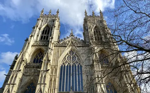 The majestic York Minster, a Gothic cathedral, stands tall against a bright blue sky with scattered clouds. Bare tree branches frame the intricate stonework and towering spires.
