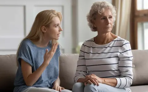 Junge Frau spricht vorwurfsvoll zu ihrer Mutter, die abgewandt sitzt. Die Mutter beleidigt die erwachsene Tochter.