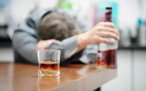 Zaniedbany mężczyzna, prawdopodobnie po eksmisji alkoholika z domu, leży na stole obok szklanki z whisky i butelki.