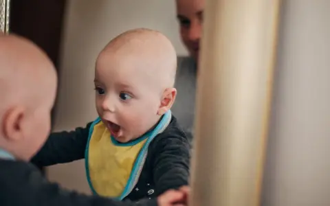 Ein Baby mit gelbem Lätzchen schaut überrascht in den Spiegel. Es ist fasziniert von seinem Spiegelbild, ein Moment, wann sagen Kinder "Ich".