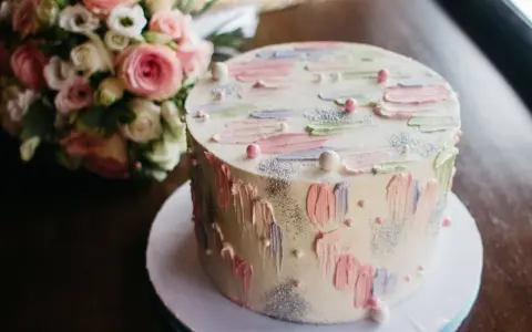 Oto tort na 20 osób, ważący około 2 kg, ozdobiony pastelowymi pociągnięciami pędzla i perełkami. Obok bukiet różowych kwiatów.