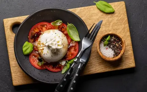 Pyszna burrata z pomidorami, polana oliwą, udekorowana bazylią. Obok miseczka z solą i pieprzem.