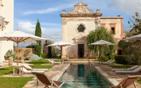 Piscina e lettini in una masseria per bambini in Puglia, con un antico edificio sullo sfondo.