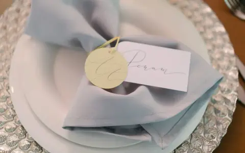 Elegancka winietka z imieniem "Peram" na jasnoszarym obrusie, ozdobiona okrągłym, żółtym tagiem.
