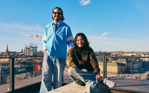 Två glada personer på en takterrass med utsikt över Stockholm, en solig dag. I bakgrunden syns stadens silhuett och en glimt av pelago slussen.