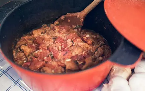En röd gryta med en mustig toscansk tomatsoppa, en träsked rör om i den väldoftande grytan. Bredvid ligger vitlök.