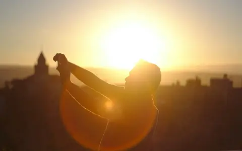 Silhouette d'une personne prenant une photo contre jour avec son téléphone, le soleil éclatant à l'horizon derrière des bâtiments.