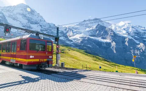 Treno rosso e giallo in stazione, con imponenti montagne innevate sullo sfondo. Un viaggio in treno tra le vette.