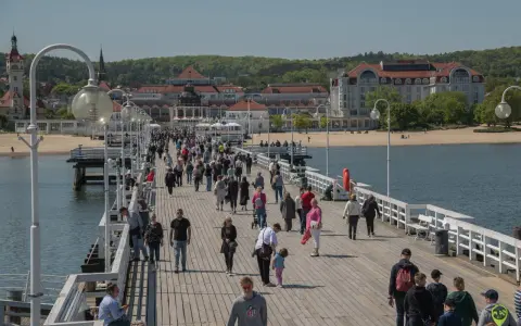 Tłum ludzi spaceruje po molo w Sopocie, jednym z nietypowych atrakcji Gdańska. W tle widać plażę i zabytkowe budynki.