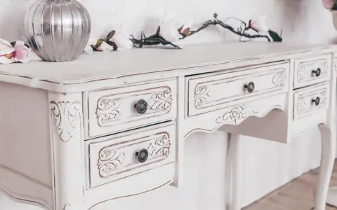 Toeletta in legno con **tecnica shabby a due colori**, decorata con intagli floreali e pomelli antichi. Un vaso d'argento e fiori rosa completano la scena.