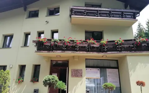 Jasnozielony hotel na błoniach z balkonem pełnym czerwonych kwiatów.
