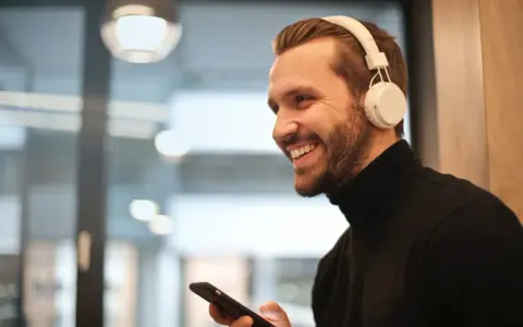 Uśmiechnięty mężczyzna w białych słuchawkach na uszach trzyma telefon, być może właśnie uczy się, jak sparować słuchawki z telefonem.