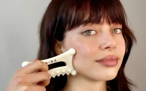 A woman with bangs and green eyes performs a facial fascia massage using a white gua sha tool.