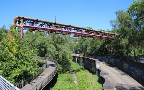Alte Rohrleitungen der Erzbahntrasse Bochum ragen über üppiges Grün. Ein geschwungener Fußweg schlängelt sich durch die Natur.
