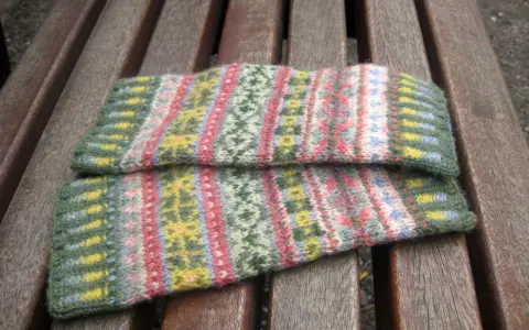 Två handledsvärmare i fair isle stickning med mönster i grönt, rosa, gult och blått ligger på en träbänk.