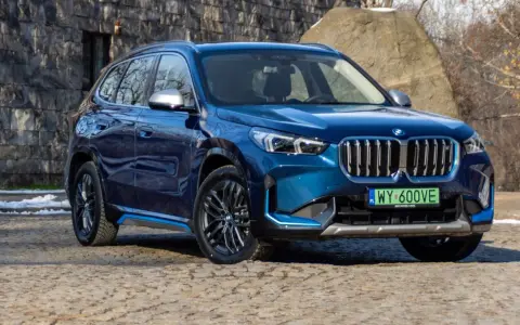 Niebieskie BMW iX1 stoi na kamienistej nawierzchni przed kamienną ścianą.