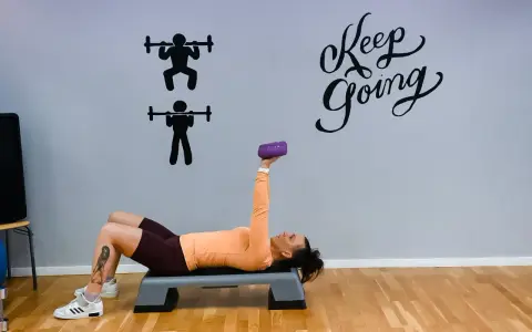 Kvinna tränar med en lila hantel på en stepbräda. Väggen har texten "Keep Going" och silhuetter av styrkelyft.