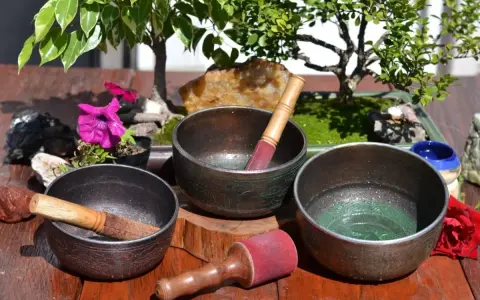Tres cuencos tibetanos para limpiar piedras, con mazos de madera y detalles ornamentales, junto a un bonsái y flores.