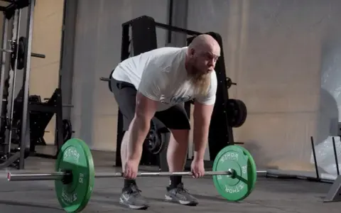 Man med skägg utför raka marklyft med hantlar i ett gym.