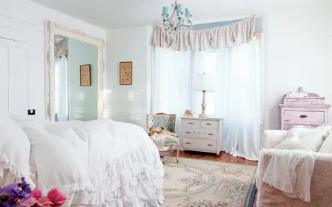 Dormitorio con cama blanca y volantes, espejo grande, cómoda antigua y sillón. Estilo shabby chic.