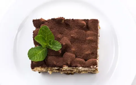 Pyszne tiramisu bez alkoholu, posypane kakao i udekorowane listkami mięty, na białym talerzu.