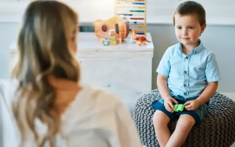 Chłopiec siedzi na pufie naprzeciwko psychoterapeutki, w tle zabawki.