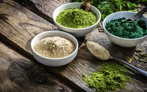 Drei Schalen mit Pulver auf Holz: hellbraun, leuchtend grün und dunkelgrün. Spirulina gesund und voller Nährstoffe.