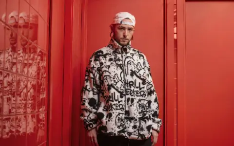El cantante Kaze, con gorra blanca y chaqueta polar con grafitis, posa en un pasillo rojo vibrante.
