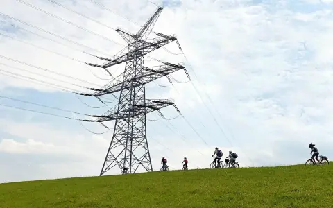 Grupa rowerzystów przemierza zielone wzgórze pod potężną konstrukcją linii 110 kV.
