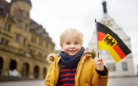 Uśmiechnięty chłopiec z blond włosami trzyma flagę Niemiec. W tle widać zabytkowe budynki, przypominające te, które można spotkać w niemieckich miasteczkach, gdzie znajdują się szkoły.