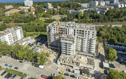 Budowa bloku mieszkalnego w Kołobrzegu. Widok z lotu ptaka na plac budowy, dźwig, samochody i okoliczne budynki. Ceny mieszkań Kołobrzeg mogą być różne.