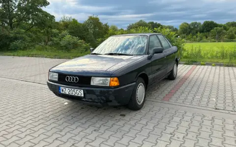 Ciemny Audi 80, klasyk z lat 90., idealny dla szukających aut do 5 tys. zł. Zaparkowany na kostce brukowej, otoczony zielenią.