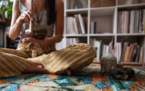 Mujer en posición de loto, realizando un ritual de limpieza energética con un cuenco tibetano. A su lado, una cabeza de Buda y un incienso humeante.