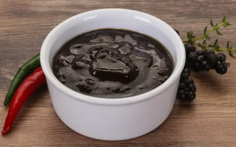 Gęsty, ciemny sos pieprzowy w białej miseczce, obok czerwona i zielona papryczka chili oraz gałązka jeżyn.