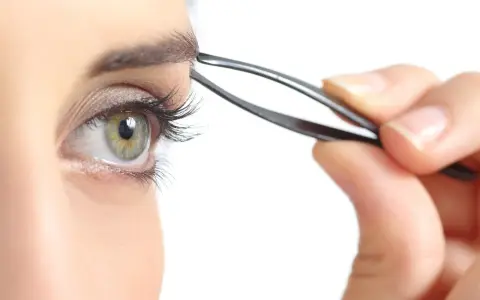 Une main utilise une pince à épiler pour parfaire l'épilation des sourcils d'un œil vert aux longs cils.