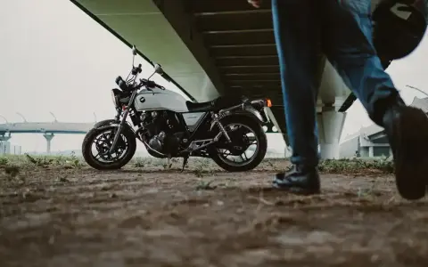 Biały motocykl klasyczny, kategoria A2, pod mostem. Obok przechodzi motocyklista z kaskiem.