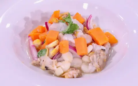 Delicioso ceviche de corvina con trozos de camote, choclo, cebolla roja y culantro, servido en un plato blanco.