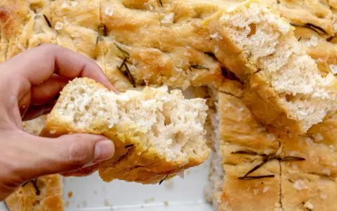 Mano rompiendo un trozo de focaccia casera con romero y sal gruesa. ¡Una delicia de esta focaccia receta!