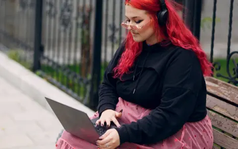 Młoda babista z ognistymi włosami i słuchawkami na uszach pracuje na laptopie, siedząc na ławce w parku.