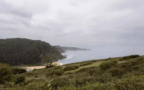 Paisaje costero con acantilados verdes y mar gris. Ideal para senderismo en Villaviciosa, con una playa dorada y una casa blanca.