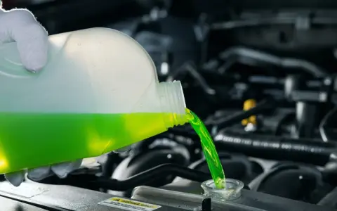 A gloved hand pours green antifreeze and coolant into a car's engine.