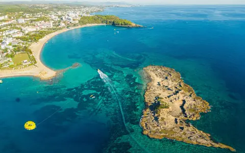 Widok z lotu ptaka na wybrzeże z hotelami, plażą i turkusową wodą. Widać łódź, parasailing i skaliste wysepki, które są świetnymi okurcalar atrakcje.