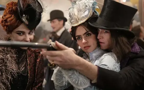 Una donna con un cappello piumato punta una pistola, protetta da un uomo con un cilindro. Un'altra donna con un cappello decorato sorride. Un momento intenso da "Dracula a love tale".
