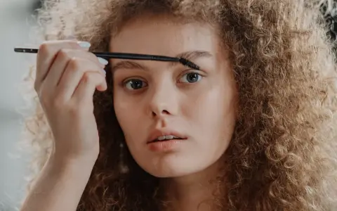 Une jeune femme aux cheveux bouclés applique un produit sur ses sourcils décolorés avec une brosse.