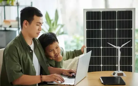 Père et fils étudient l'énergie solaire et éolienne sur un ordinateur portable, un panneau solaire et une éolienne à proximité. Un projet d'installation cacsi sans consuel.