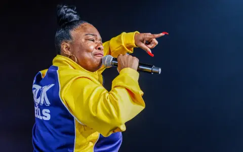 Roxanne Shanté w żółtej kurtce z niebieskimi wstawkami śpiewa do mikrofonu, wskazując palcem.