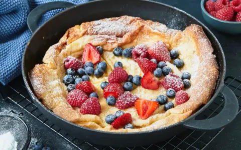 Ein goldbrauner Dutch Baby mit Puderzucker und frischen Beeren, serviert in einer gusseisernen Pfanne.