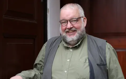Ein freundlicher evangelischer Pfarrer mit Brille und Bart lächelt in die Kamera, vor einer dunklen Holztür stehend.