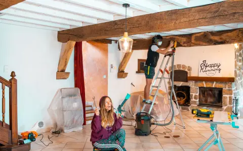 Un homme ponce une poutre en bois avec une ponceuse, tandis qu'une femme tient des outils.